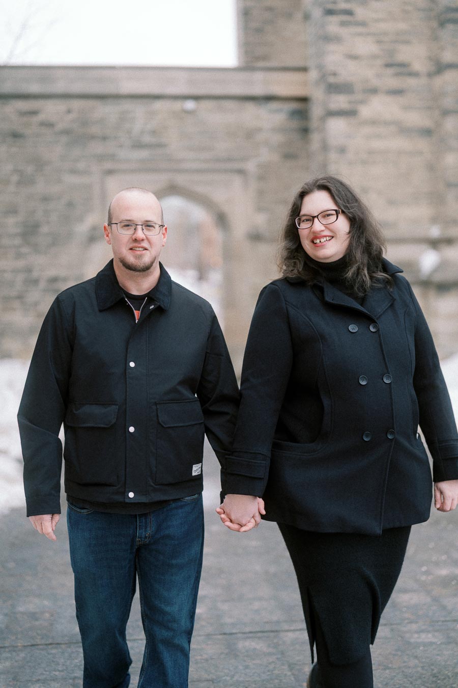 A couple holding hands and walking on a snowy pathway in front of a historic stone building. Both are wearing winter coats and glasses, smiling at the camera.