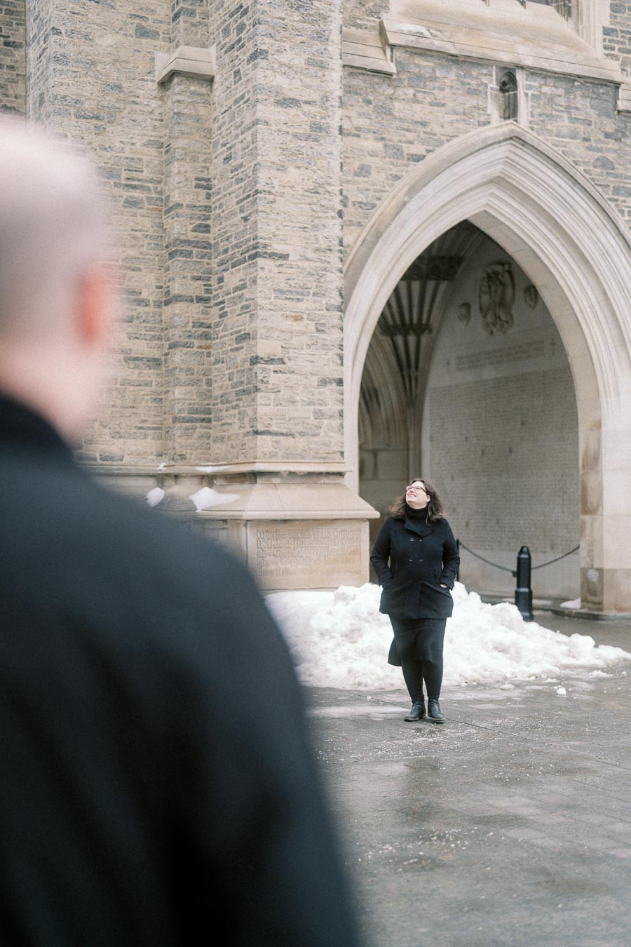 A woman in a black coat stands outside a historic stone building with arched architecture on a snowy day, while a blurred person in the foreground observes from a distance.