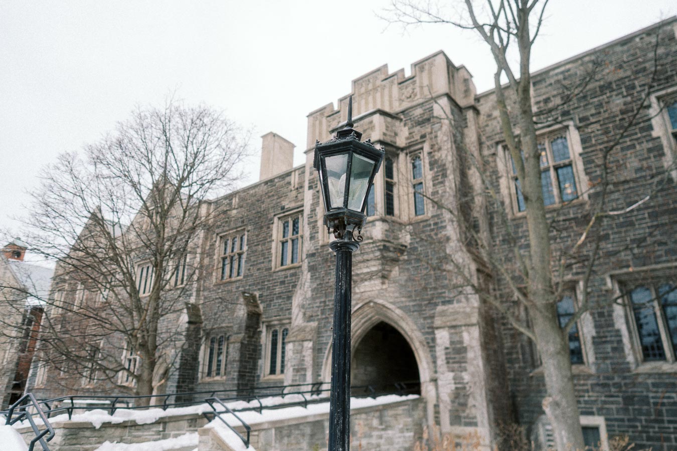 Historic Gothic-style building with stone facade and arched entrance in winter, featuring a classic black lamppost in the foreground against a backdrop of bare trees and snow.