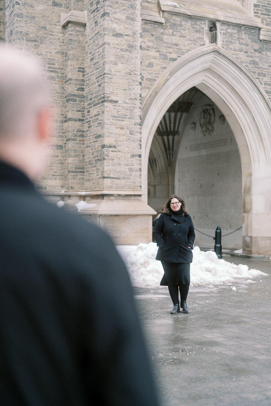 A person standing in front of a historic stone building with an arched entrance, surrounded by snow on a winter day.