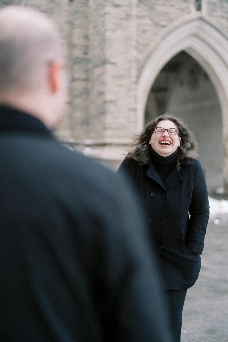 A woman in a black coat joyfully laughing outdoors in front of a stone building with an archway, facing a blurred figure in the foreground.