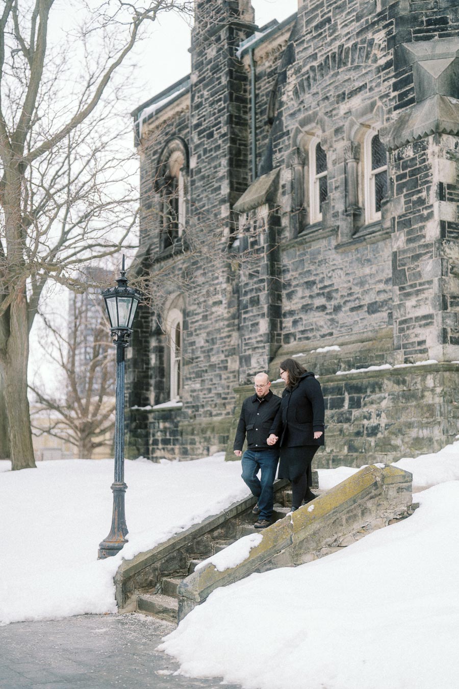 Couple walking hand in hand down snowy stone steps next to a historic stone building, with bare trees and a vintage street lamp in the winter setting.