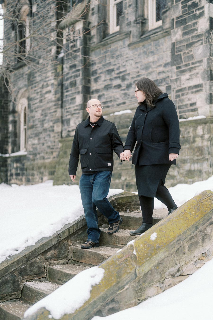 Couple walking hand in hand on snowy steps beside a stone building, wearing winter coats and looking at each other.
