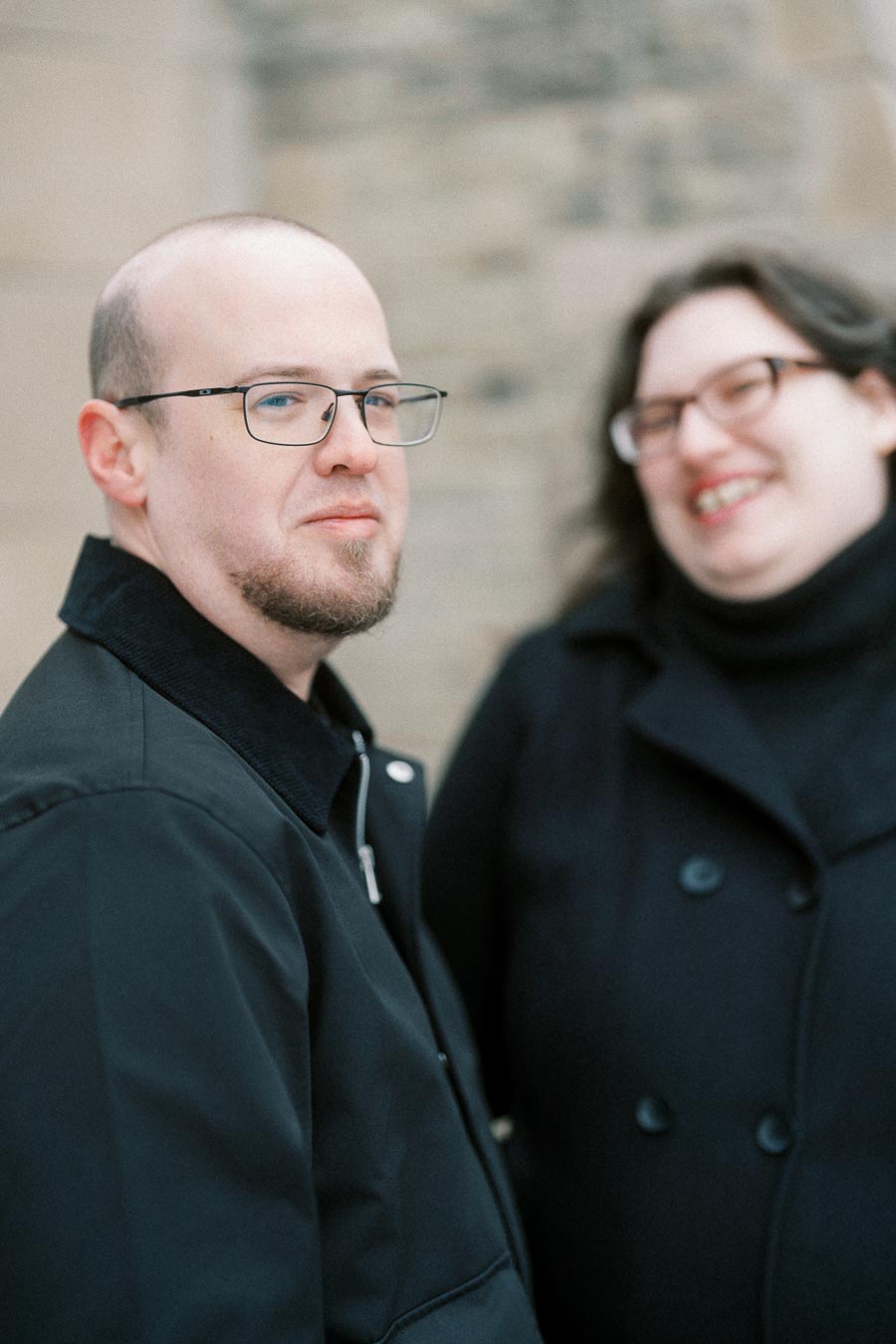 A man and woman in black coats standing outdoors, smiling and looking towards the camera, with a blurred stone wall background.