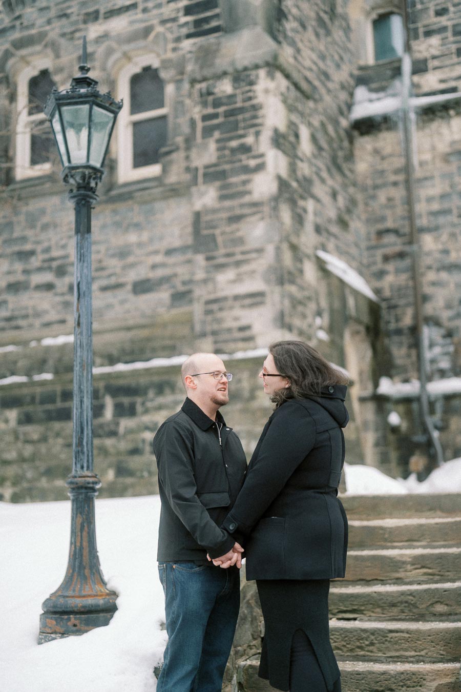 A couple in winter clothing holding hands and smiling near a historic stone building with a vintage lamp post, surrounded by snow.