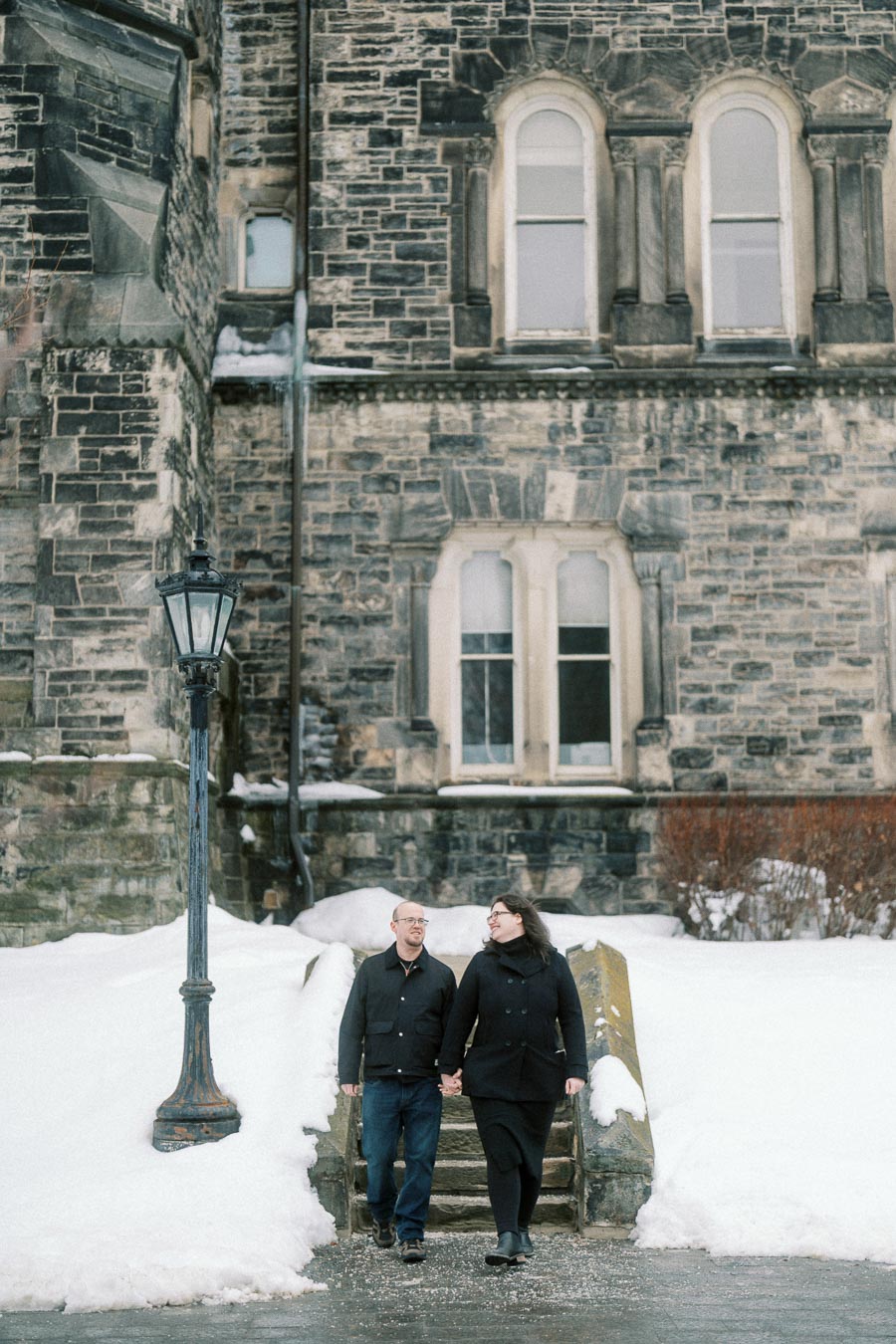 A couple holding hands and walking down snowy steps in front of a historic stone building with arched windows and a vintage lamp post in winter.