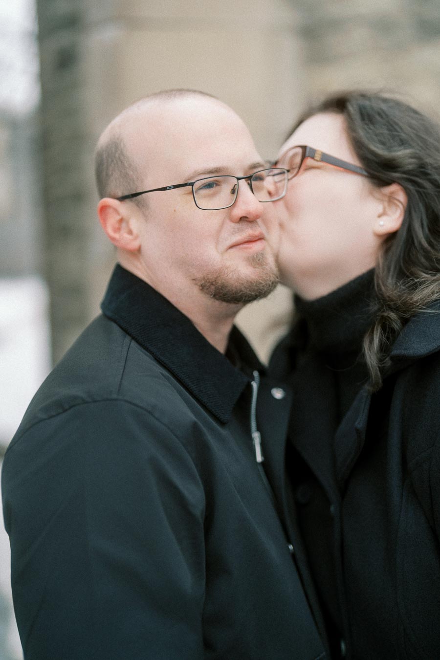 A woman playfully kisses a man on the cheek outdoors, both dressed warmly in black coats.