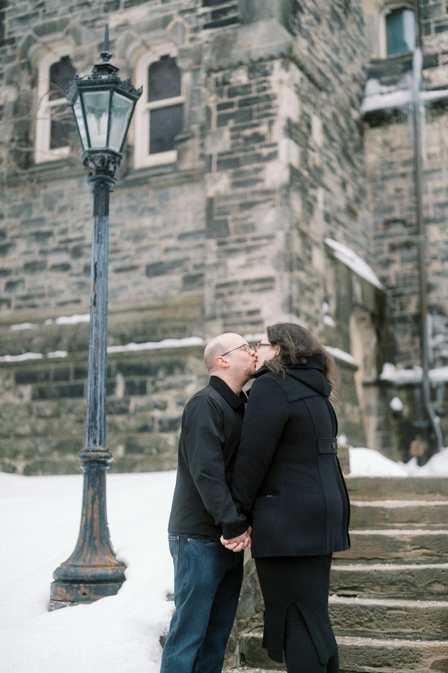 A couple kisses in front of a historic stone building during winter, with snow covering the ground and a vintage street lamp nearby.