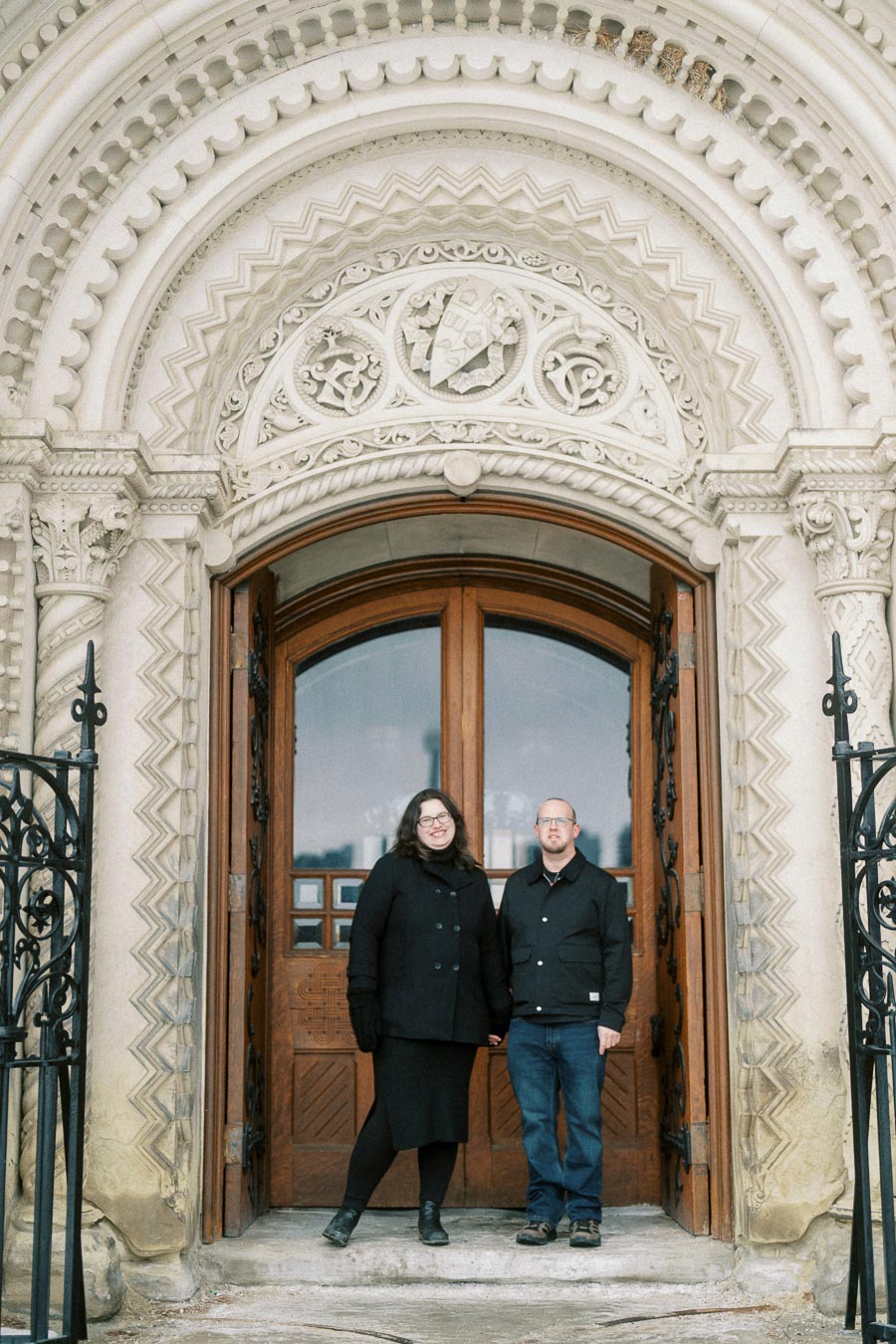 Couple standing in front of an ornate, historical building entrance, featuring intricate stone carvings and a wooden door.