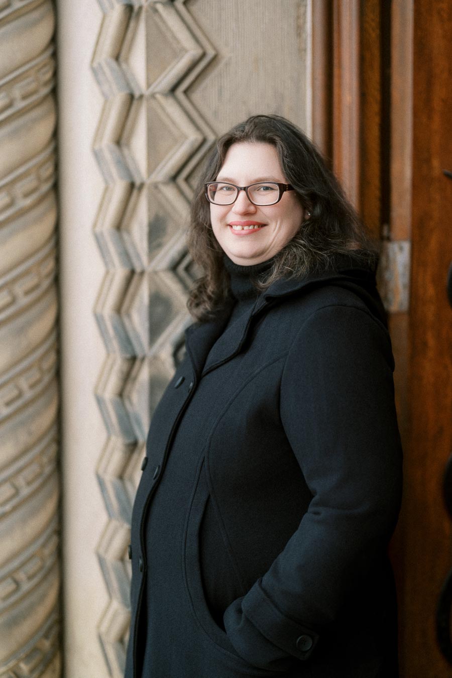 Smiling person in a dark coat standing in front of an ornate architectural wall and wooden door.