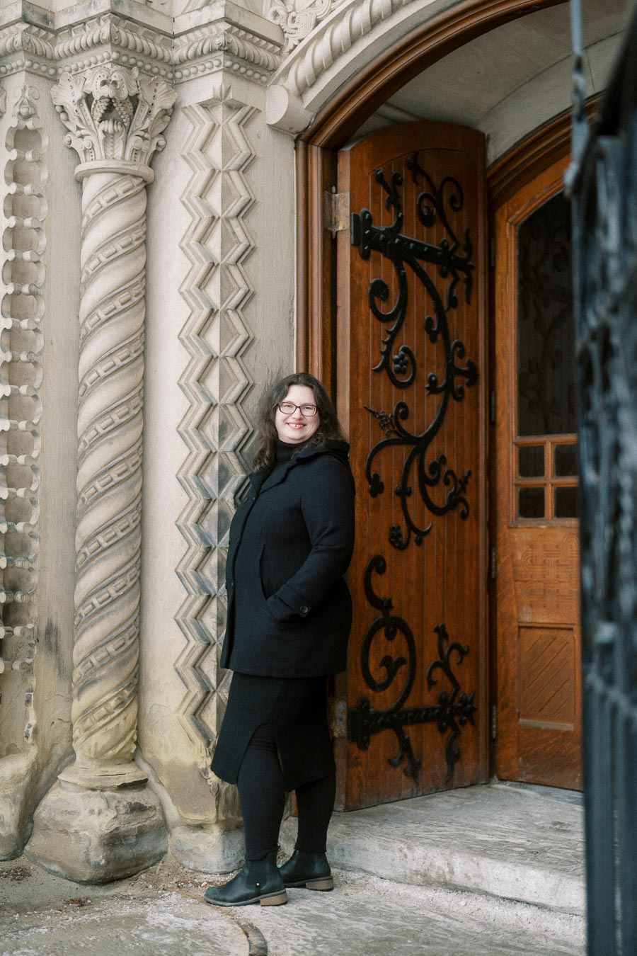 Woman in a black coat smiling near an intricately carved wooden door with ornate stone detailing.