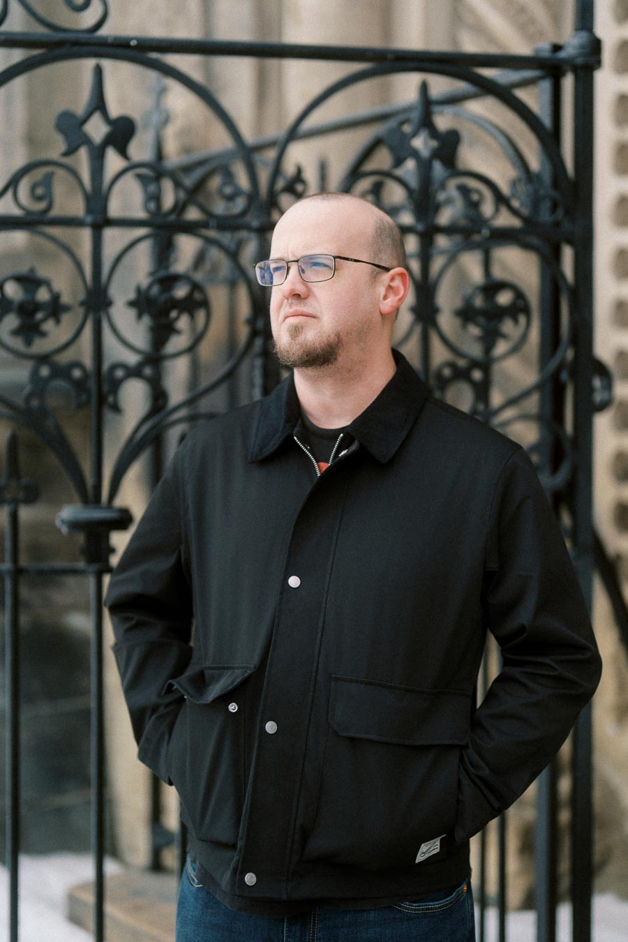 Man in a black jacket with glasses standing in front of ornate wrought iron gate, looking thoughtful.