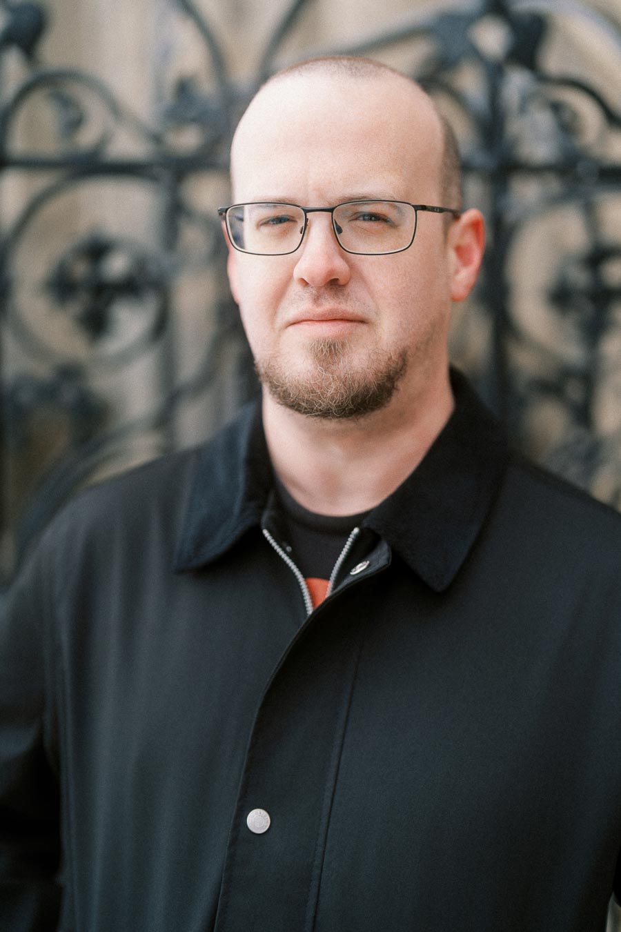 A man with glasses and a short beard, wearing a black coat, stands in front of an ornate wrought-iron fence.