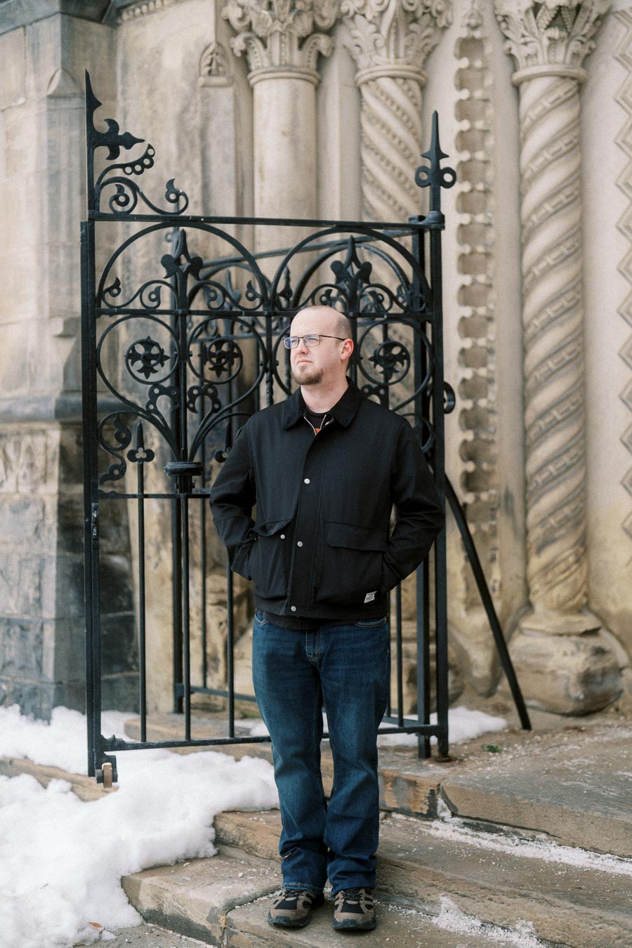 Man standing in front of ornate wrought iron gate with historic stone architecture in the background, dressed in a black jacket and blue jeans on a snowy day.