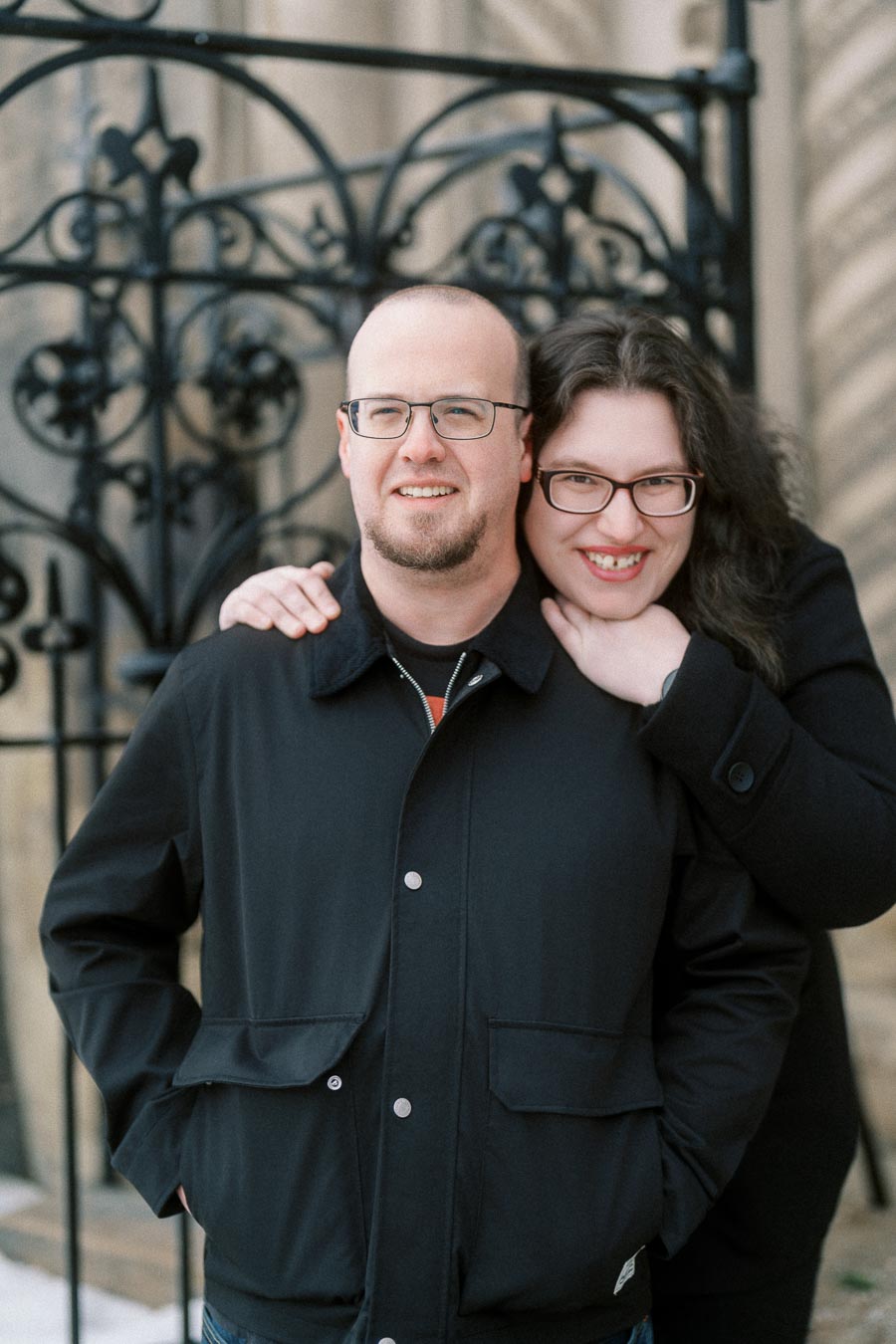 A smiling couple wearing glasses poses in front of intricate wrought ironwork, dressed warmly, indicating cooler weather.