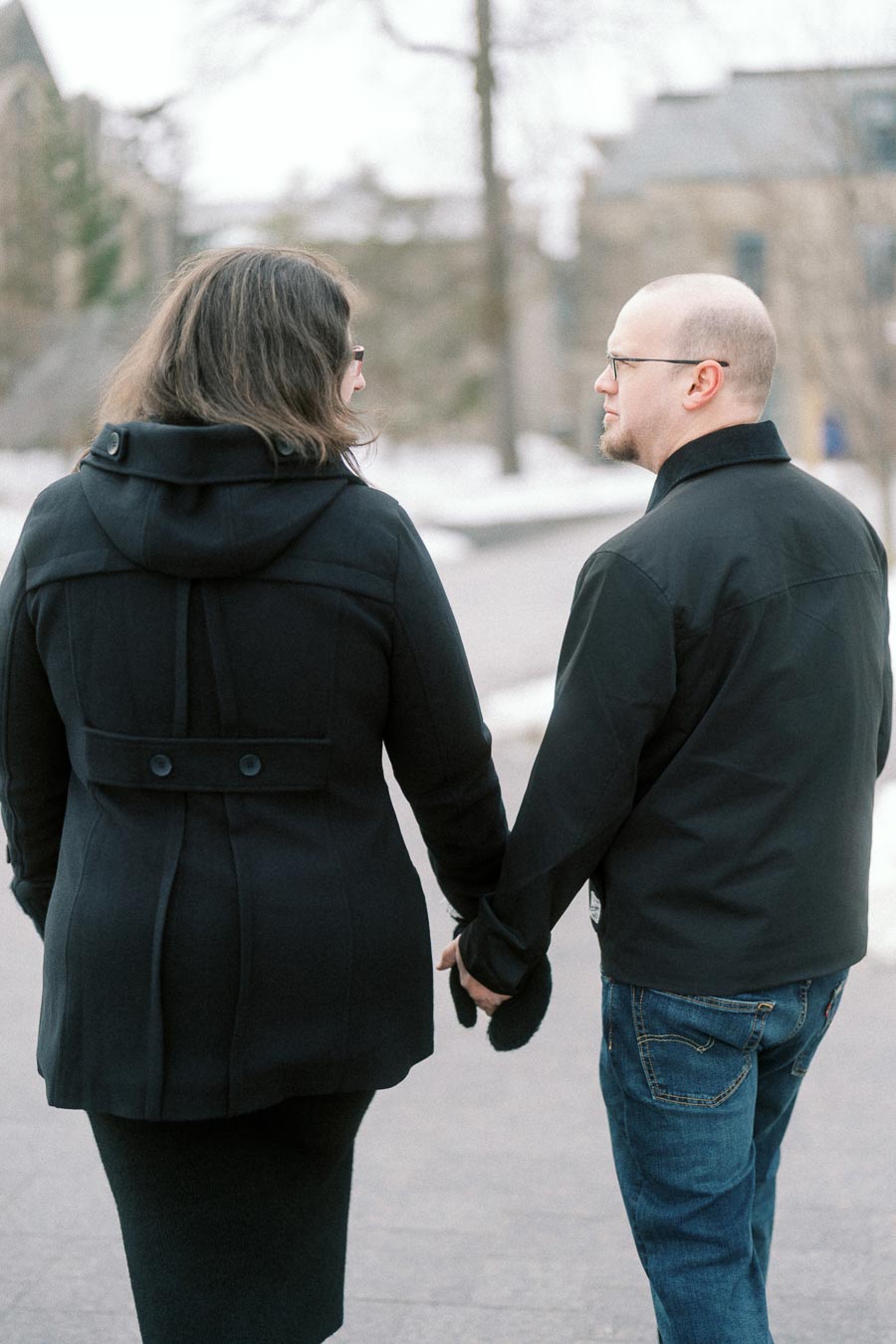A couple walking hand in hand on a snowy path, both wearing dark winter coats, with blurred trees and buildings in the background.
