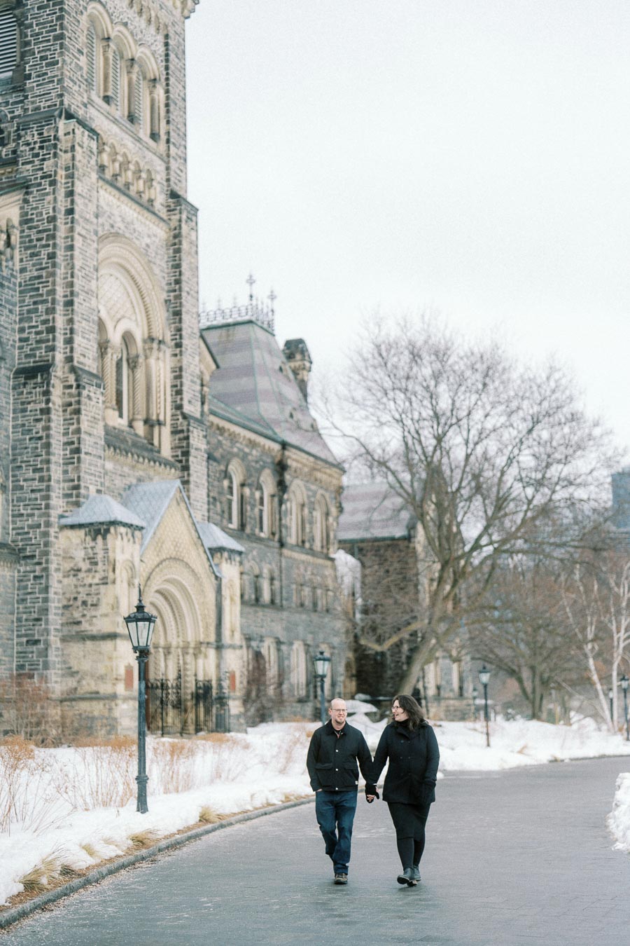 Couple walking hand in hand along a snowy pathway beside a historic stone building, showcasing a winter campus setting.