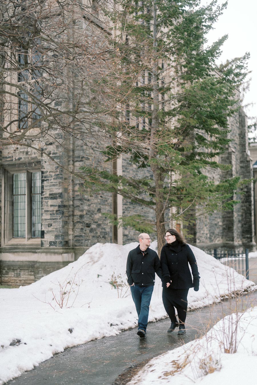 A couple walking hand in hand on a snow-covered path in front of a historic stone building, surrounded by tall trees and winter scenery.