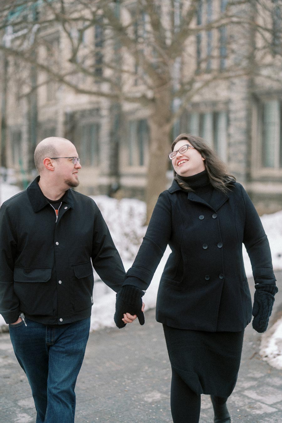 A couple dressed in winter attire, smiling and holding hands while walking on a snowy path in front of a historic building.