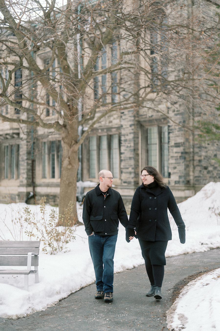 A couple walking hand in hand on a snowy path, surrounded by a picturesque winter landscape with a historic stone building in the background.