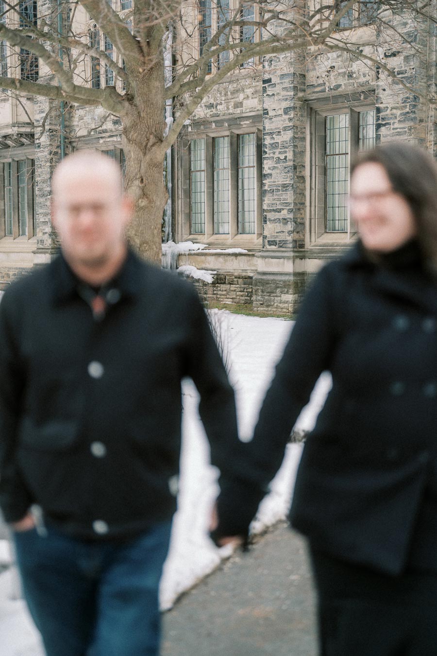 A blurred image of two people walking hand in hand in a snowy setting, with a historic stone building and leafless tree in the background.