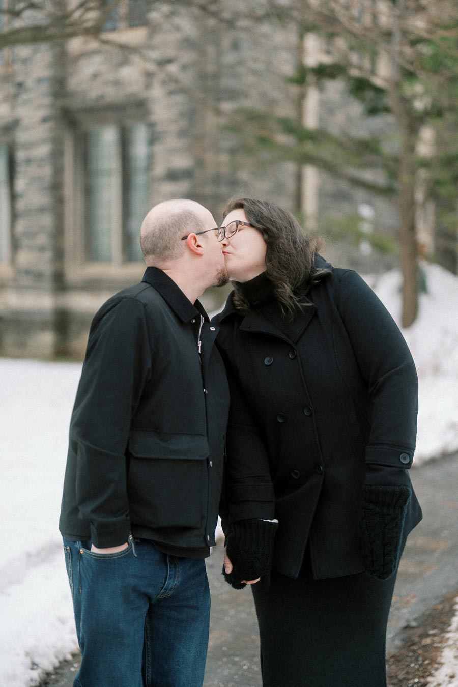 Couple in winter clothing sharing a kiss outdoors by a historic stone building, surrounded by snow-covered ground.