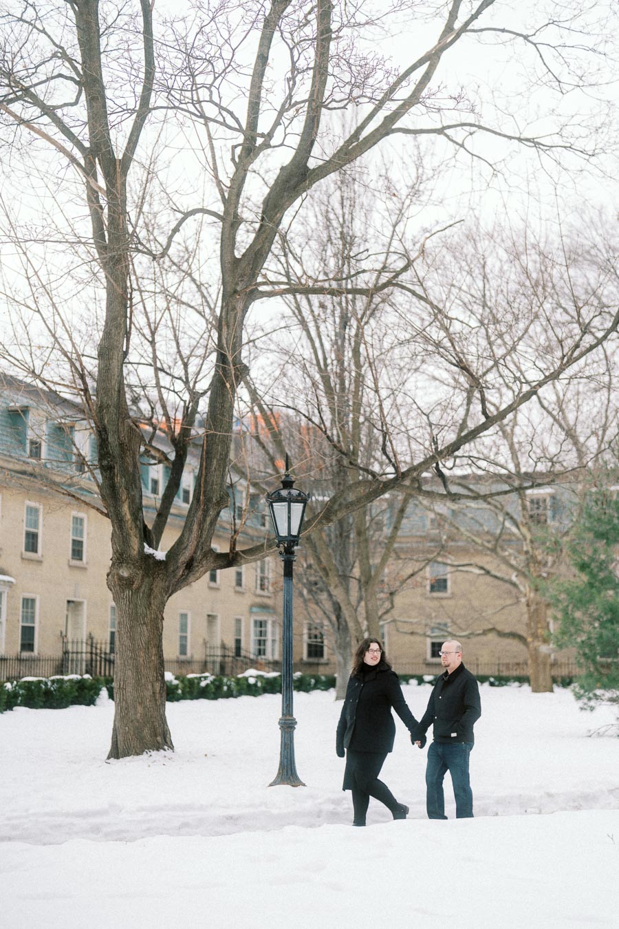 A couple holding hands and walking through a snowy park, surrounded by leafless trees and historic buildings, during winter.