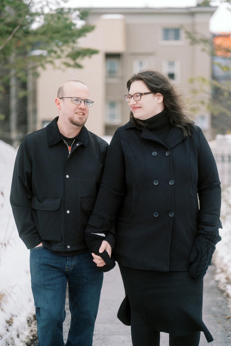 A couple walking hand in hand on a snowy pathway, wearing winter coats and gloves, with a blurred building and trees in the background.