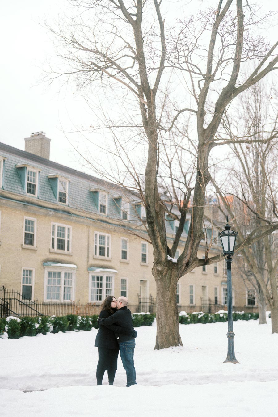 Romantic couple embraces in a snowy urban park, with historic building and bare winter tree in the background.