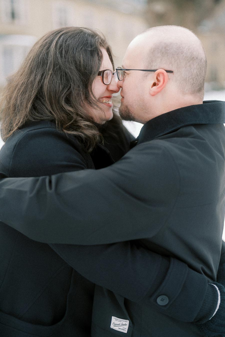 A couple dressed in black coats shares a loving embrace in a snowy, outdoor setting.