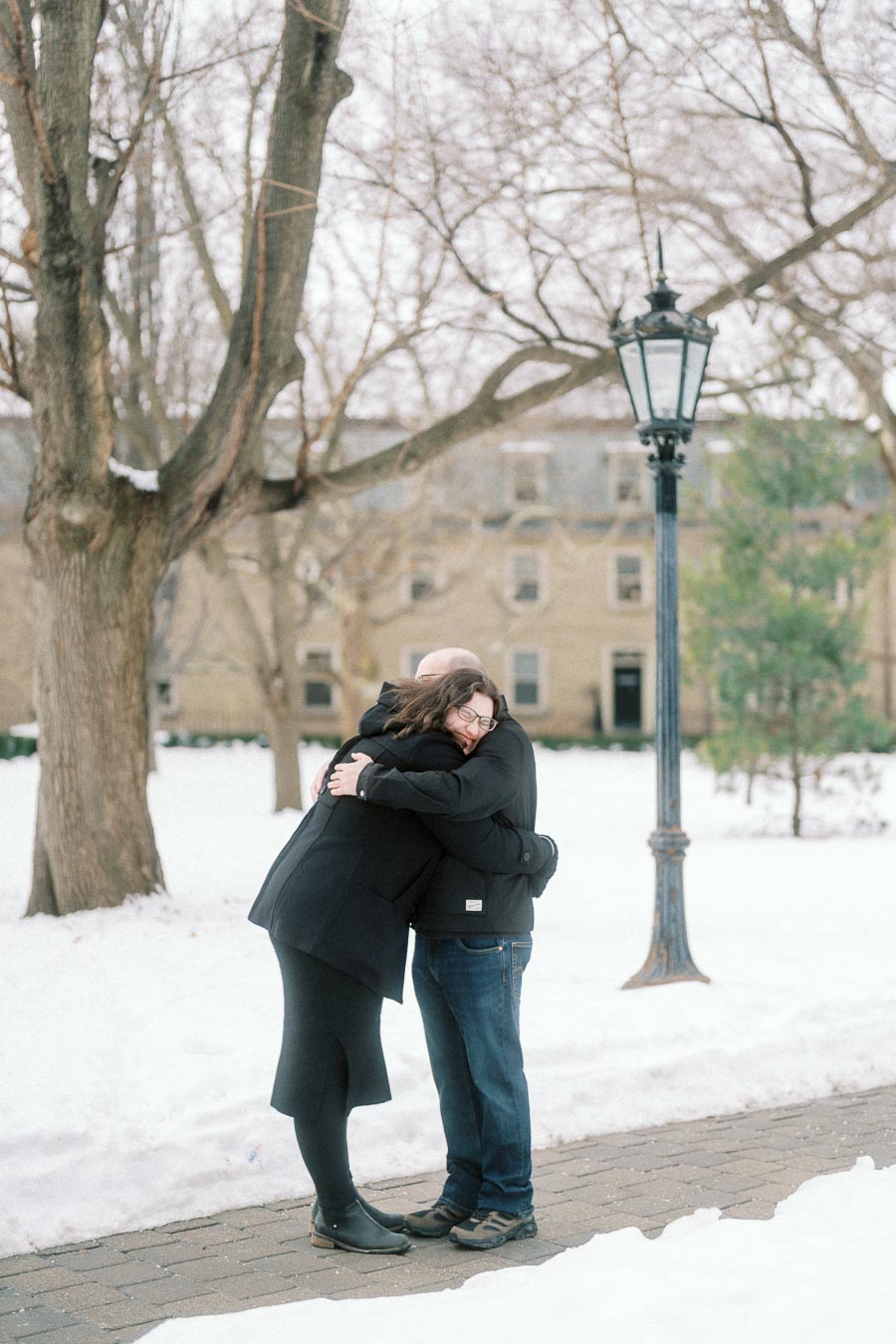 A couple warmly embracing in a snowy park, with bare trees and a historic building in the background.