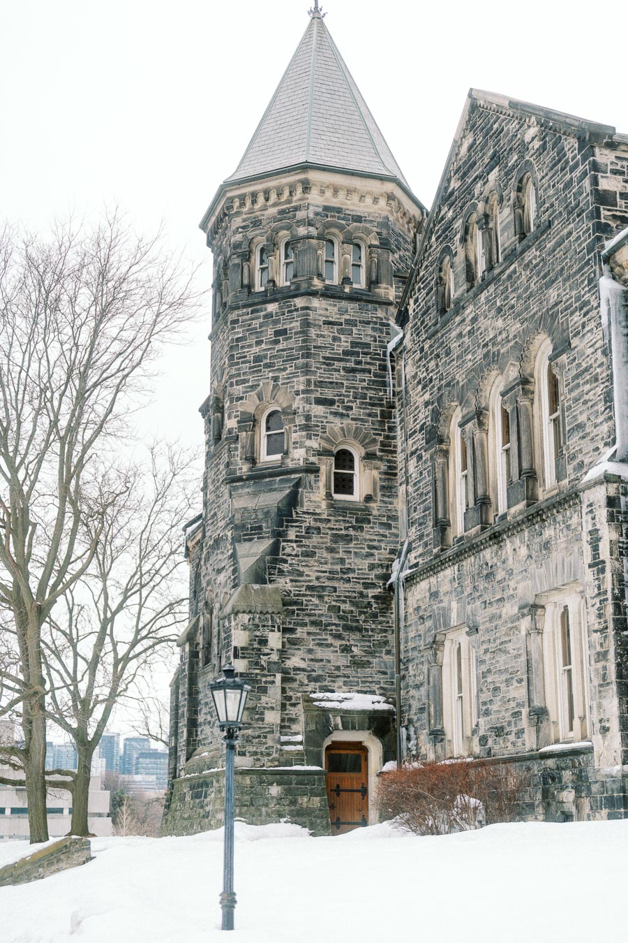 Historic stone building with a turret, surrounded by a snow-covered landscape, featuring bare trees and an old-fashioned street lamp.