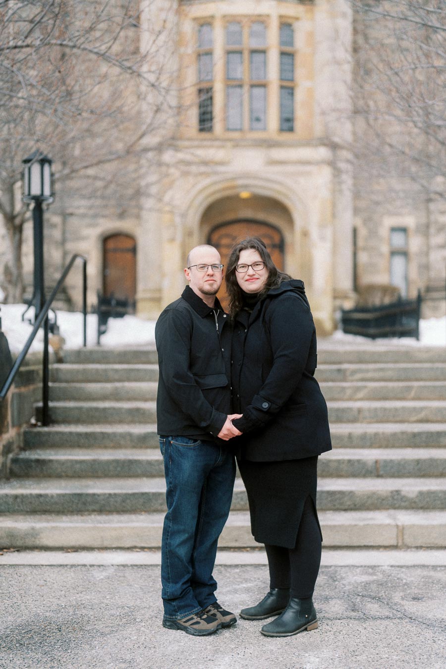 Couple in winter attire standing on stone steps in front of historic building entrance.