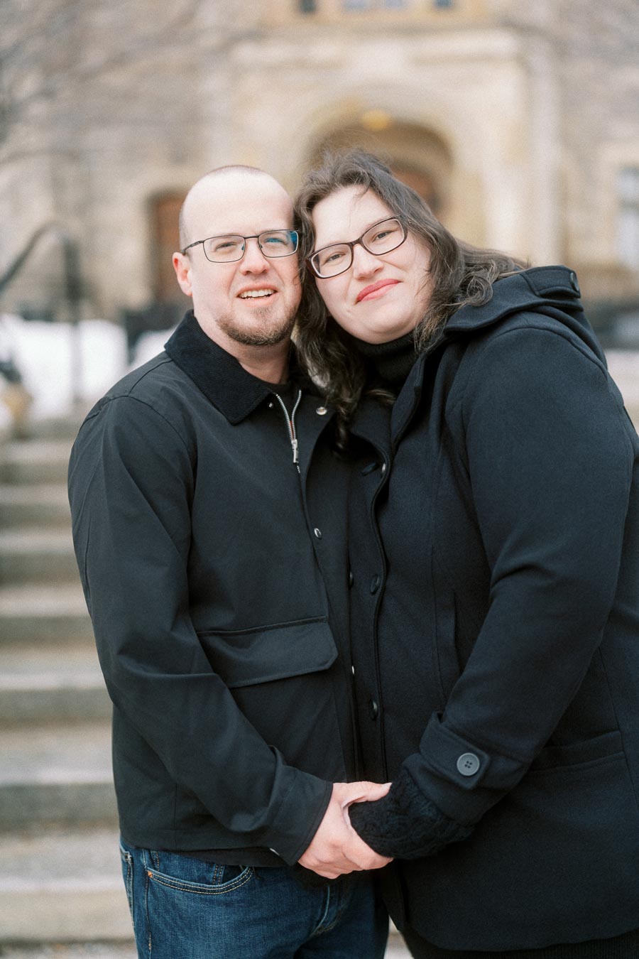 A couple standing closely together on outdoor steps, both wearing glasses and winter coats, smiling warmly at the camera, with a blurred building in the background.