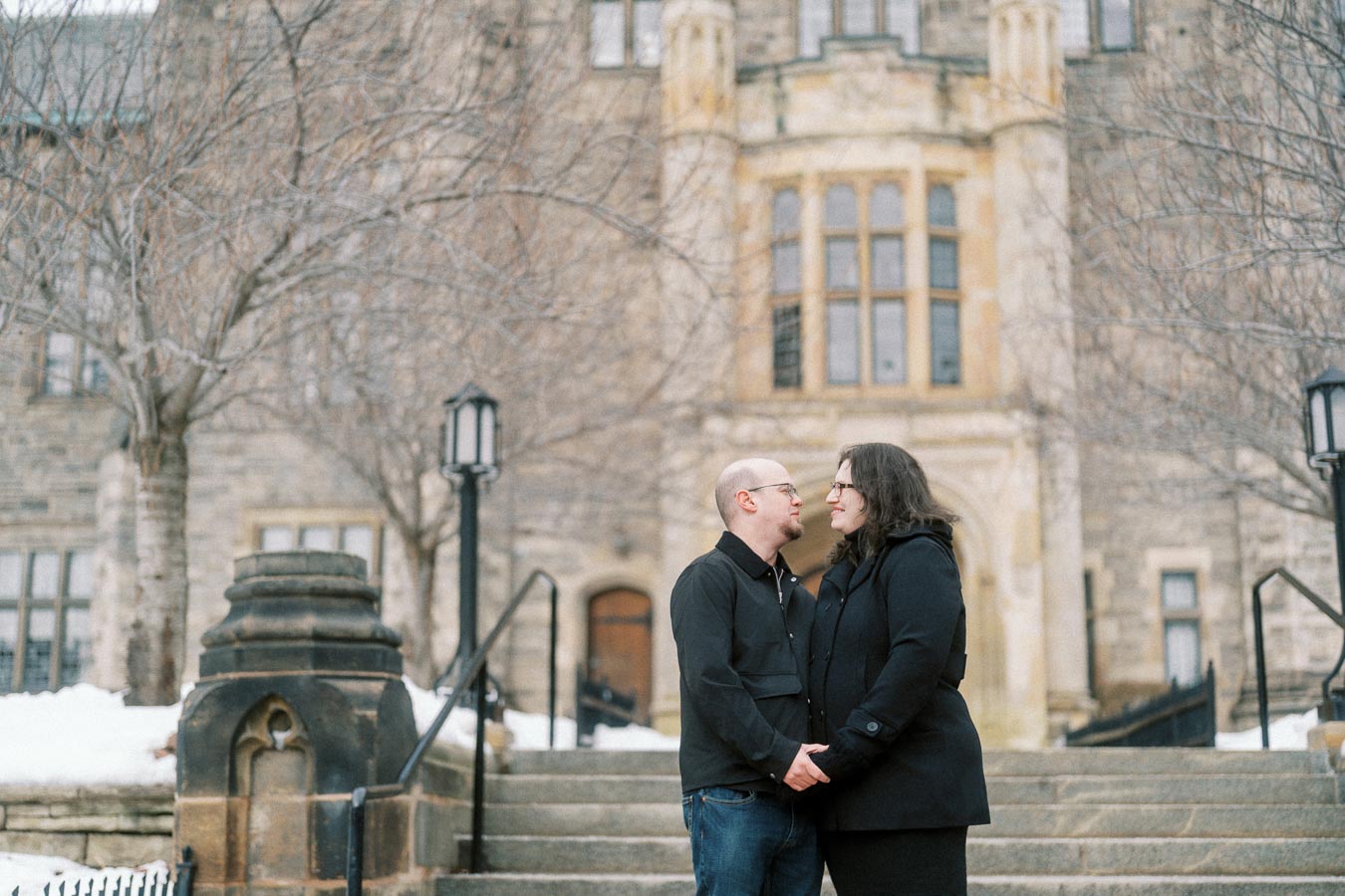 A couple holding hands and smiling at each other while standing on stone steps in front of a historic building with bare trees, winter scene.