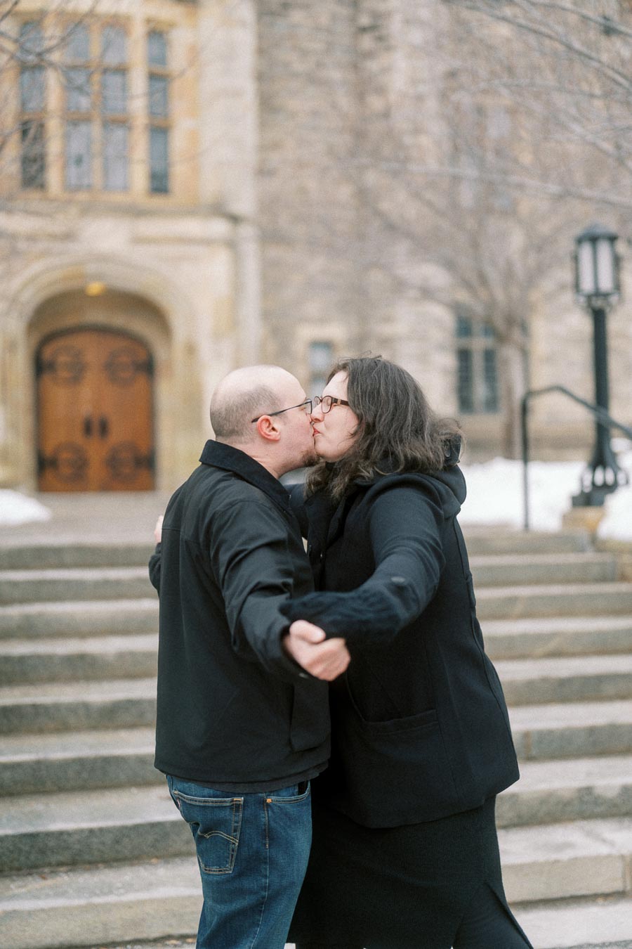 A couple warmly embracing and kissing on stone steps in front of a historic building with an ornate wooden door, capturing a romantic winter moment.