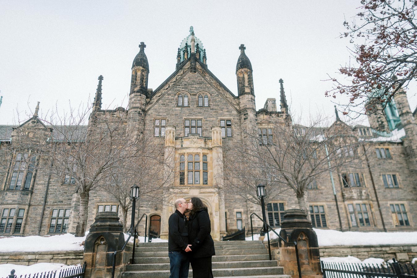 A couple sharing a kiss in front of a historic Gothic-style building during winter, with snow-covered steps and bare trees.