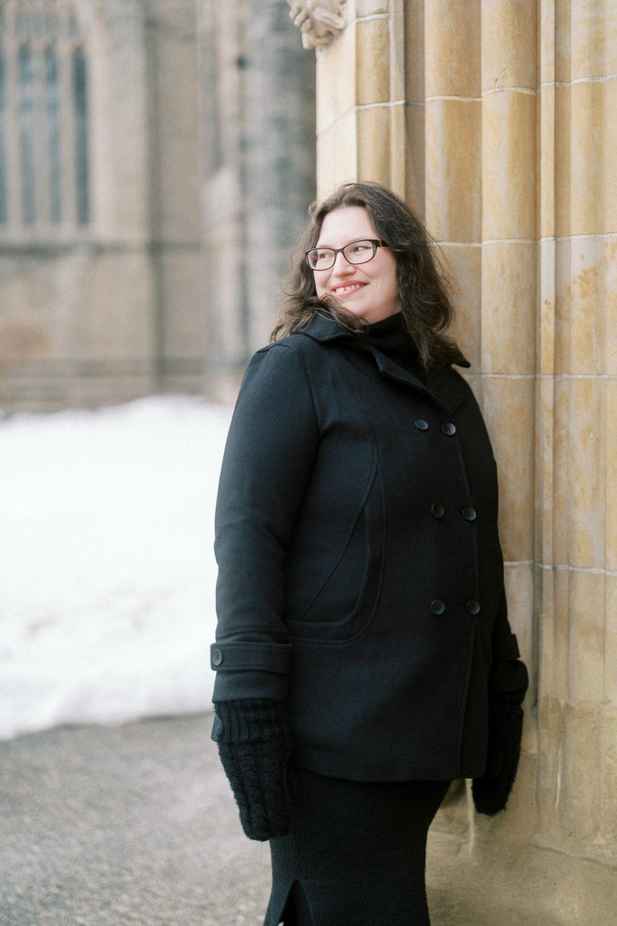 A person wearing a dark winter coat and gloves stands outdoors near a historic stone building, with snow on the ground, smiling and looking away from the camera.