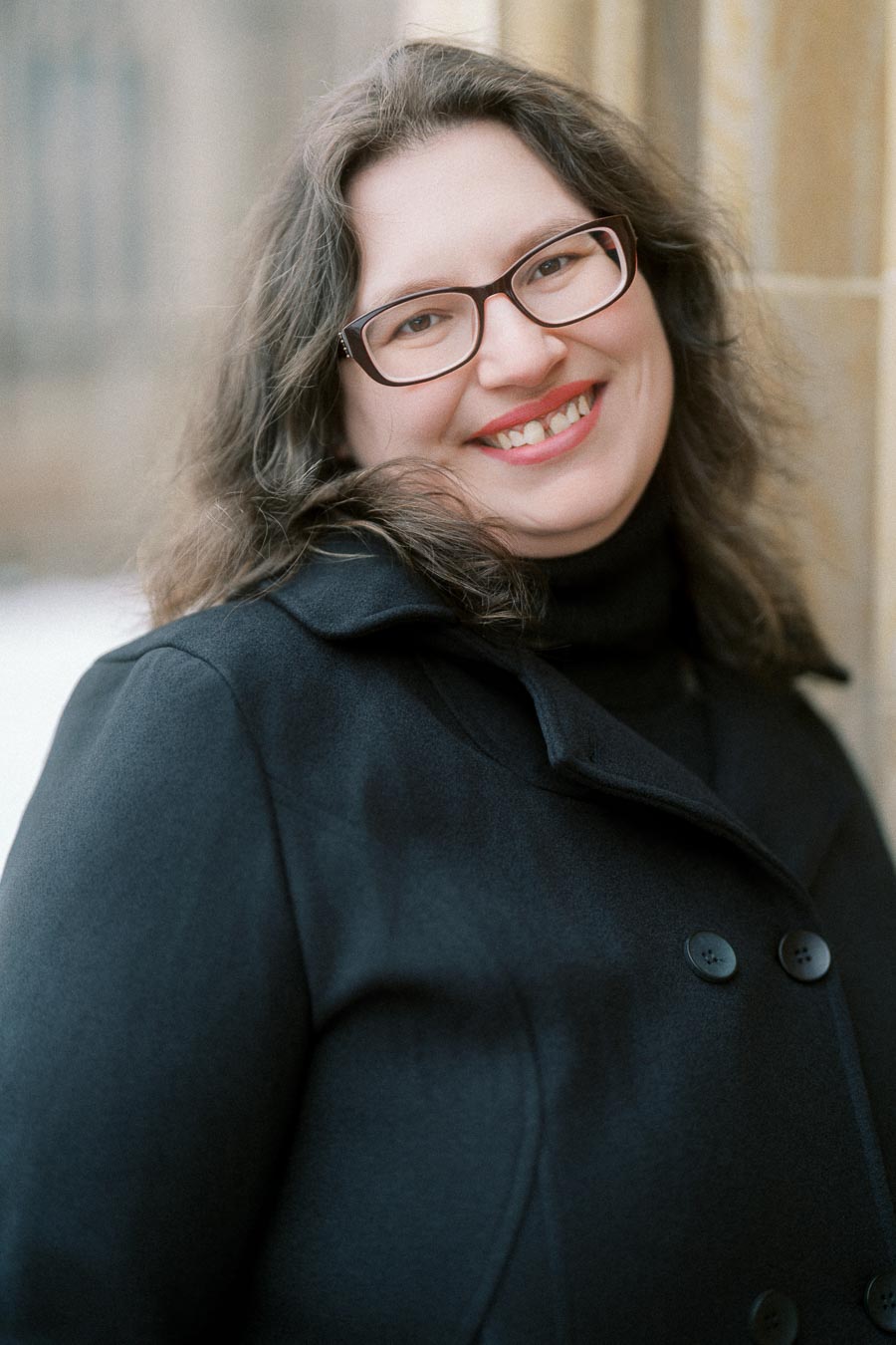 Smiling person wearing glasses and a black coat, standing outdoors near a stone column.