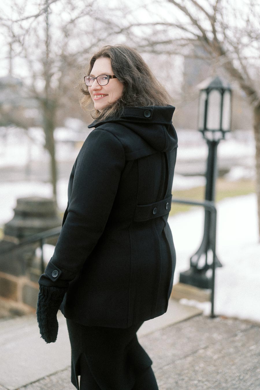 Woman in a black winter coat smiling outdoors, with snow-covered trees in the background.