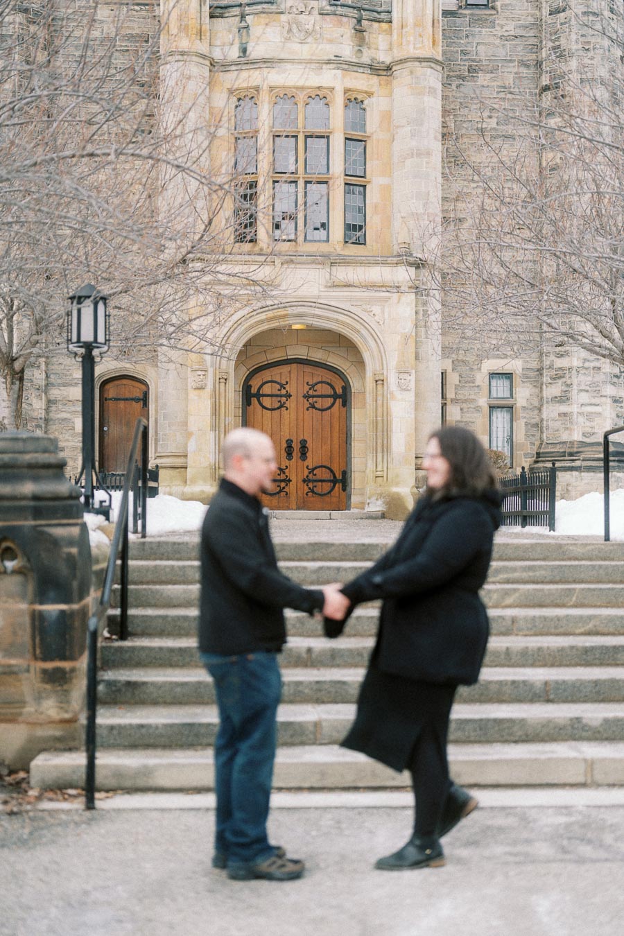 A blurred image of two people holding hands, standing on stone steps in front of an ornate, historic building with wooden doors and arched windows, surrounded by leafless trees on a winter day.