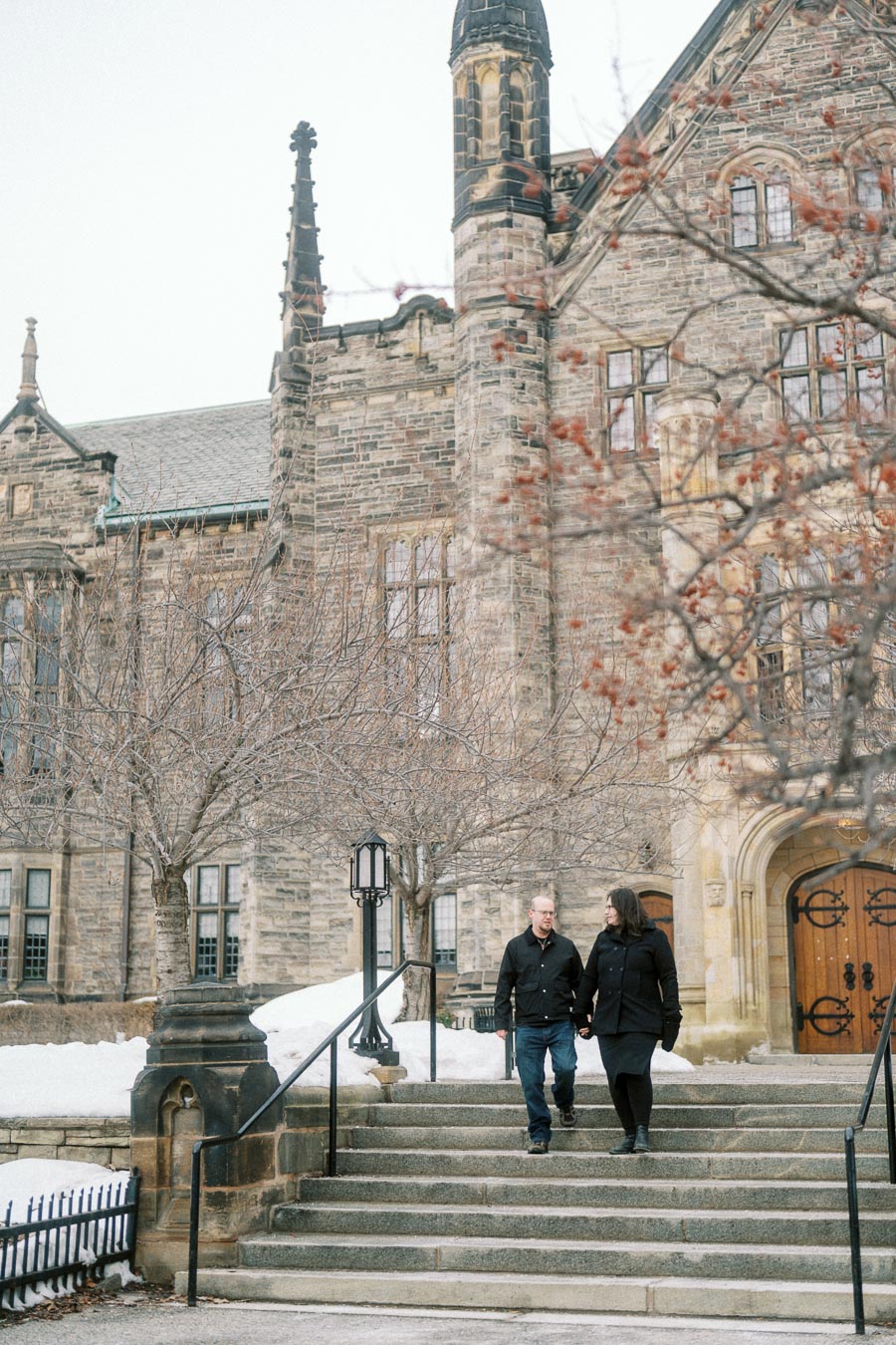 A couple walking down stone steps in front of a historic building during winter, with snow covering the ground and bare trees lining the walkway.