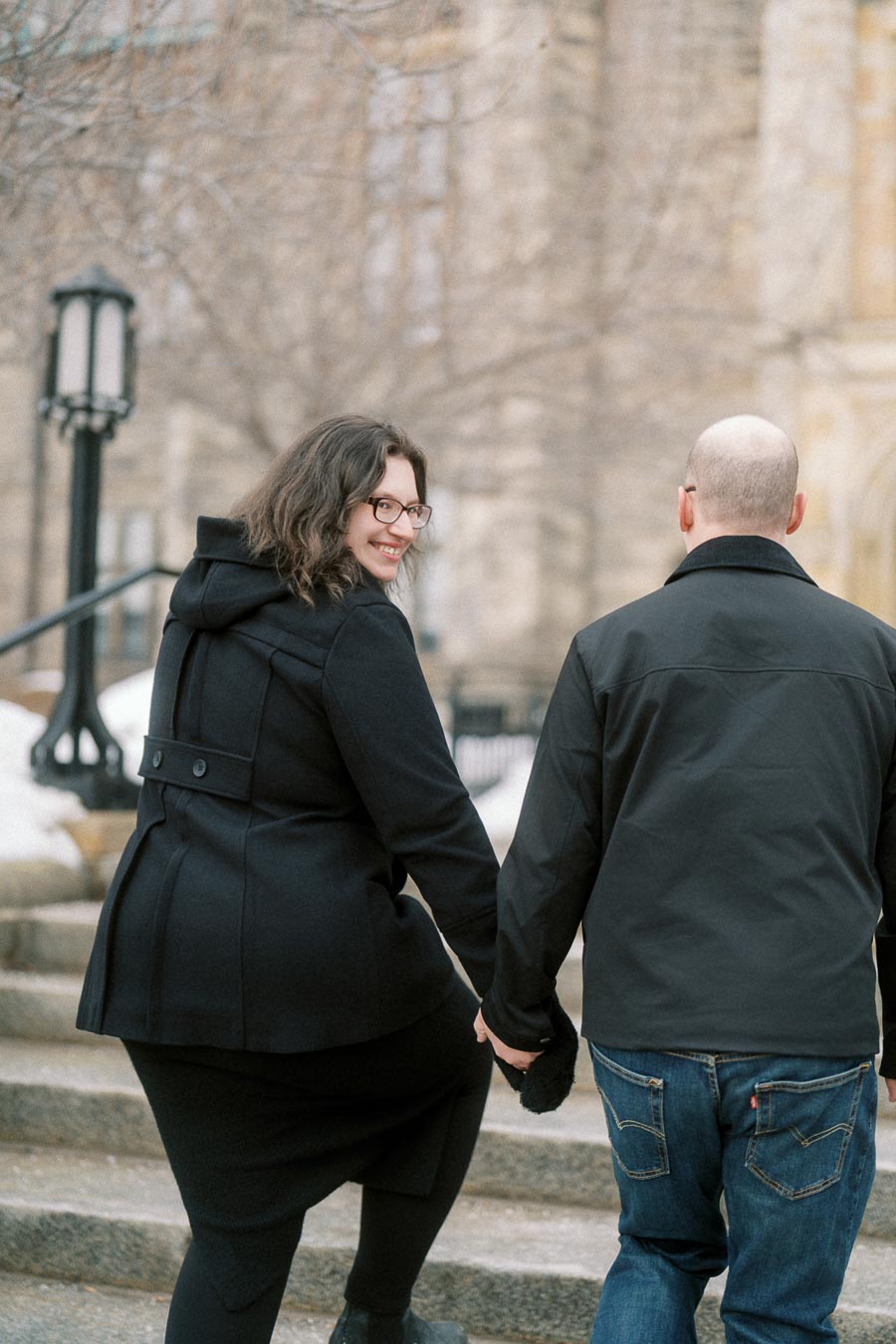 A couple in black winter coats walks hand in hand up stone steps, with trees and a historic building in the background. The woman looks back, smiling warmly.