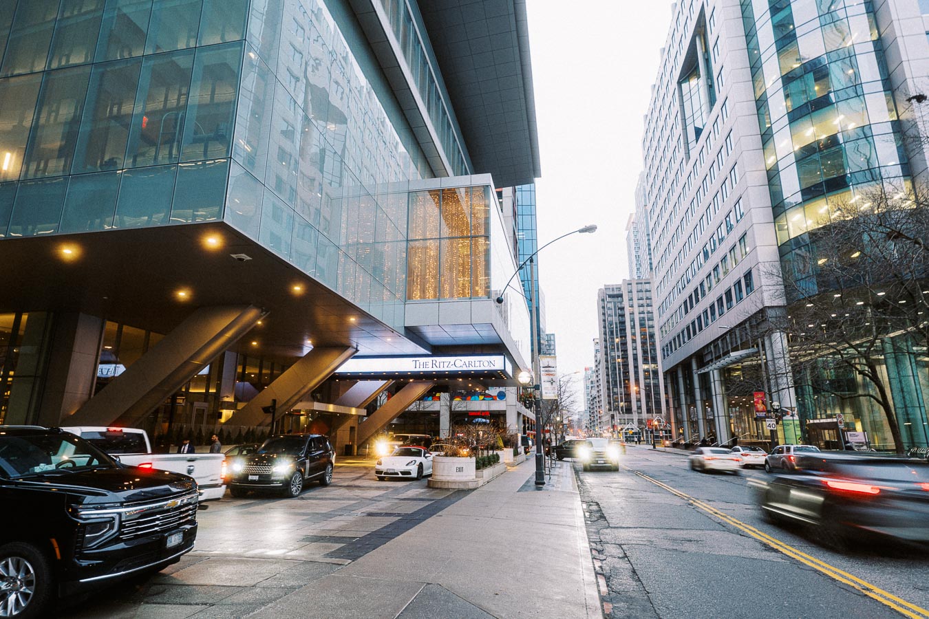 Downtown street view featuring the exterior of The Ritz-Carlton hotel with modern architecture, busy traffic, and surrounding high-rise buildings on an overcast day.