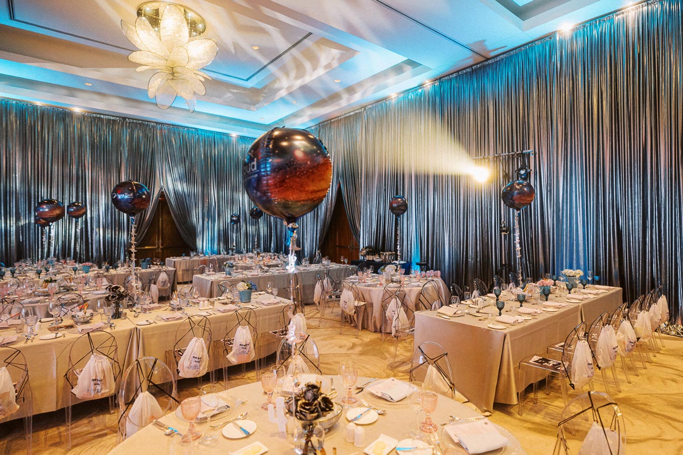 Luxurious event space with elegant table settings, featuring chic transparent chairs and metallic drapery. Large decorative balloons add a festive touch, enhancing the sophisticated atmosphere for a gala or wedding reception.