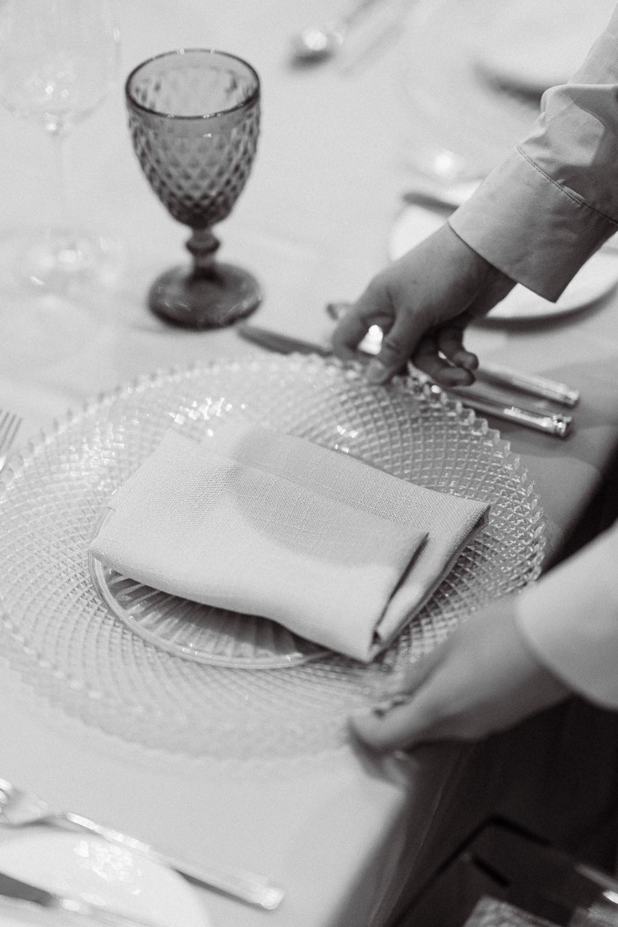 Elegant table setting with intricately patterned glass plate, neatly folded white napkin, and an ornate wine glass; person arranging the setting, highlighting fine dining or special occasion preparation.