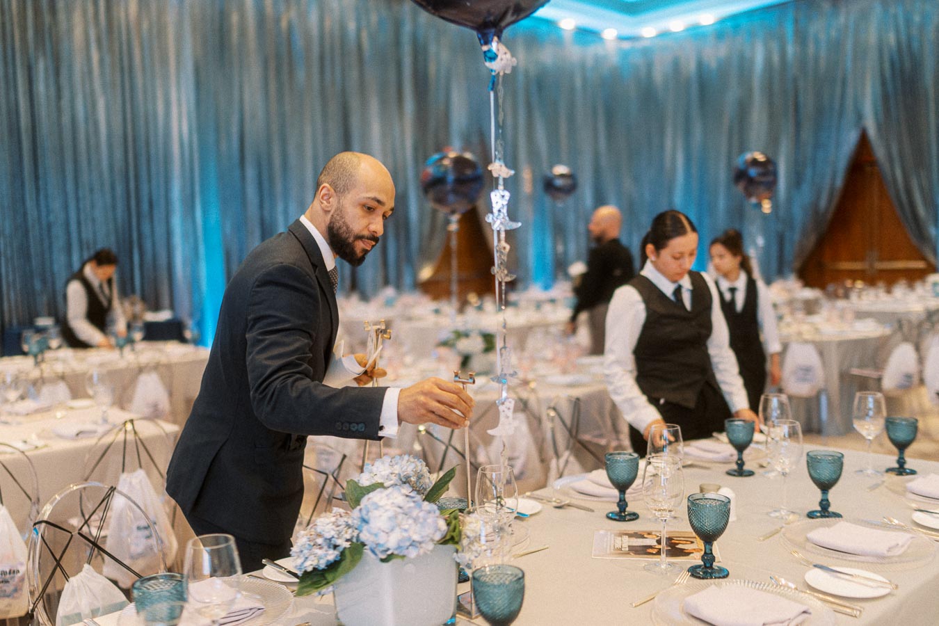 Elegant banquet hall preparation with staff setting tables, featuring blue decor elements and floral centerpieces.