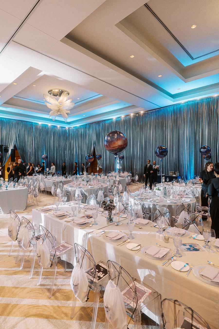 Elegant banquet hall setup with blue lighting, featuring tables with white tablecloths, clear chairs, and decorative balloons; perfect for a sophisticated corporate event or wedding reception.