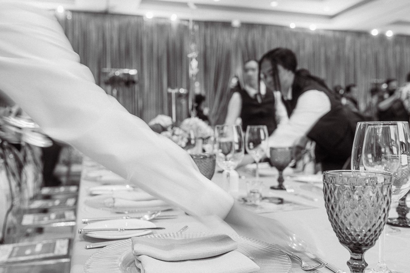 Black and white image of a formal dining table being set up by restaurant staff, featuring neatly arranged plates, cutlery, and elegant glassware, highlighting event preparation and hospitality service.