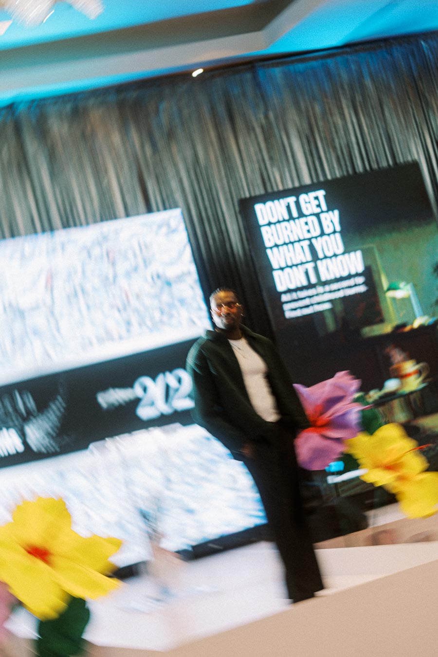 Blurry photo of a person standing on a stage with large colorful flowers and a digital display in the background, showing the year 2023 and a motivational message.